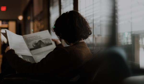 woman sitting on couch and reading the newspaper with her back facing us.