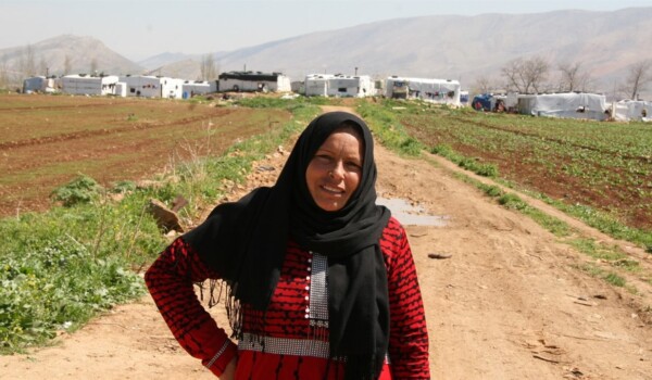 A woman in a red dress and a black burka stand outside on a field with grass on each side.