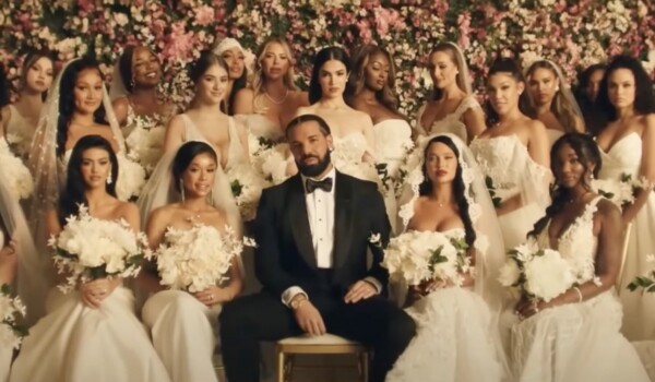 Drake in a black tux, sitting with a crowd of women in wedding dresses