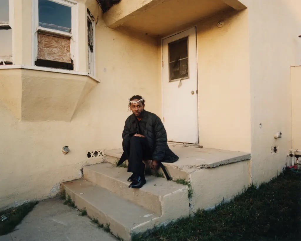 Kendrick Lamar, wearing a crown of thorns, sits on the steps outside a house