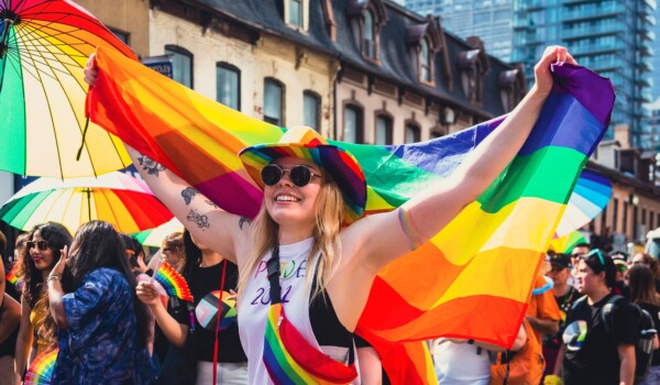 A person with long blonde hair, a rainbow cowboy hat and a rainbow fanny pack, holding a rainbow flag at the pride parade.