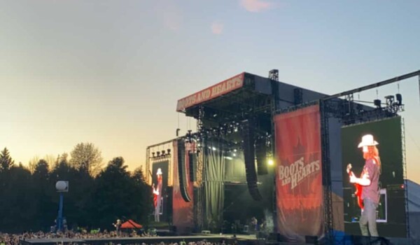 A large crowd stands in front of a stage. The sun is setting with pink clouds over the stage.