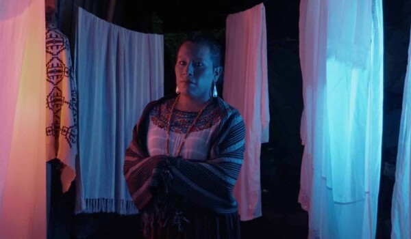 An image of Monica from the Guatemalan short film IXIM: El amor no tiene género. She sits in front of hanging white sheets lit with blue and pink lights.