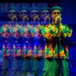 Siddartha sings into a microphone on a stand. He wears a colourful sweater, cargo shorts, and a hat. The image is overlayed with lower opacity copies of Siddartha.