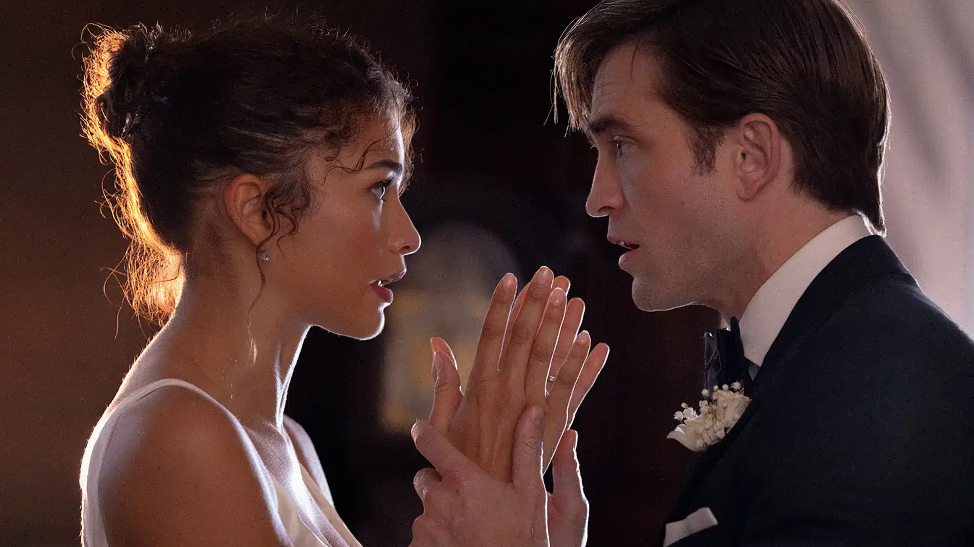 Image from the side of Charlie (Robert Pattinson) holding Emma (Zendaya) by the wrists while they look directly in eachother's eyes, dressed in wedding attire.