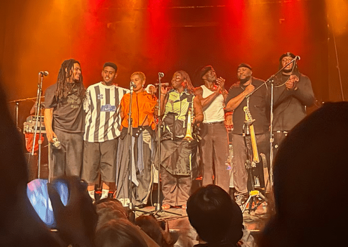 Seven members of Kokoroko in a row across the stage expressing gratitude to the audience following their concert, one member in the centre of the row has a mic and is speaking to the crowd, three on the left have their arms interlocked around eachothers' waists and shoulders, two members on the right are applauding the audience, and one member has his fist on his chest.