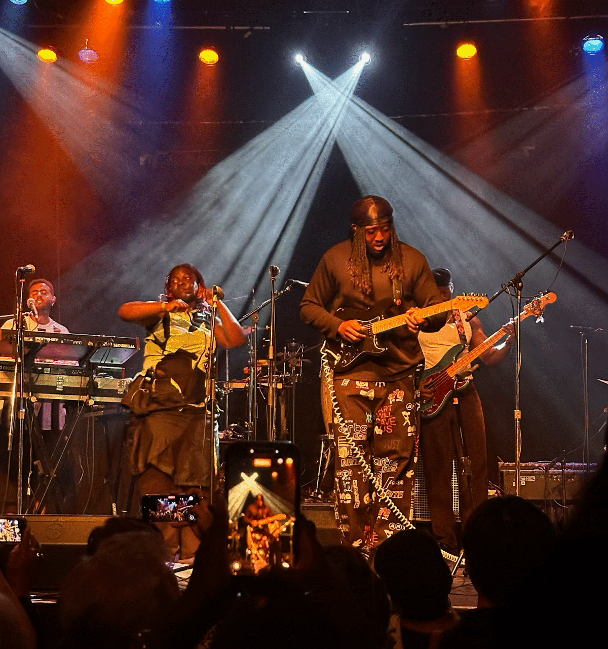 Guitarist from Kokoroko centre stage, the violinist mid-stroke and keyboard player visible in the background, with criss-crossing beams of light behind them all.